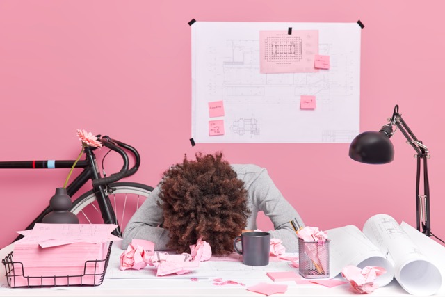 A person with curly hair sits at a cluttered desk, head down in exhaustion. The desk is covered with crumpled papers, documents, and office supplies. The background is pink with architectural plans and a bicycle visible.