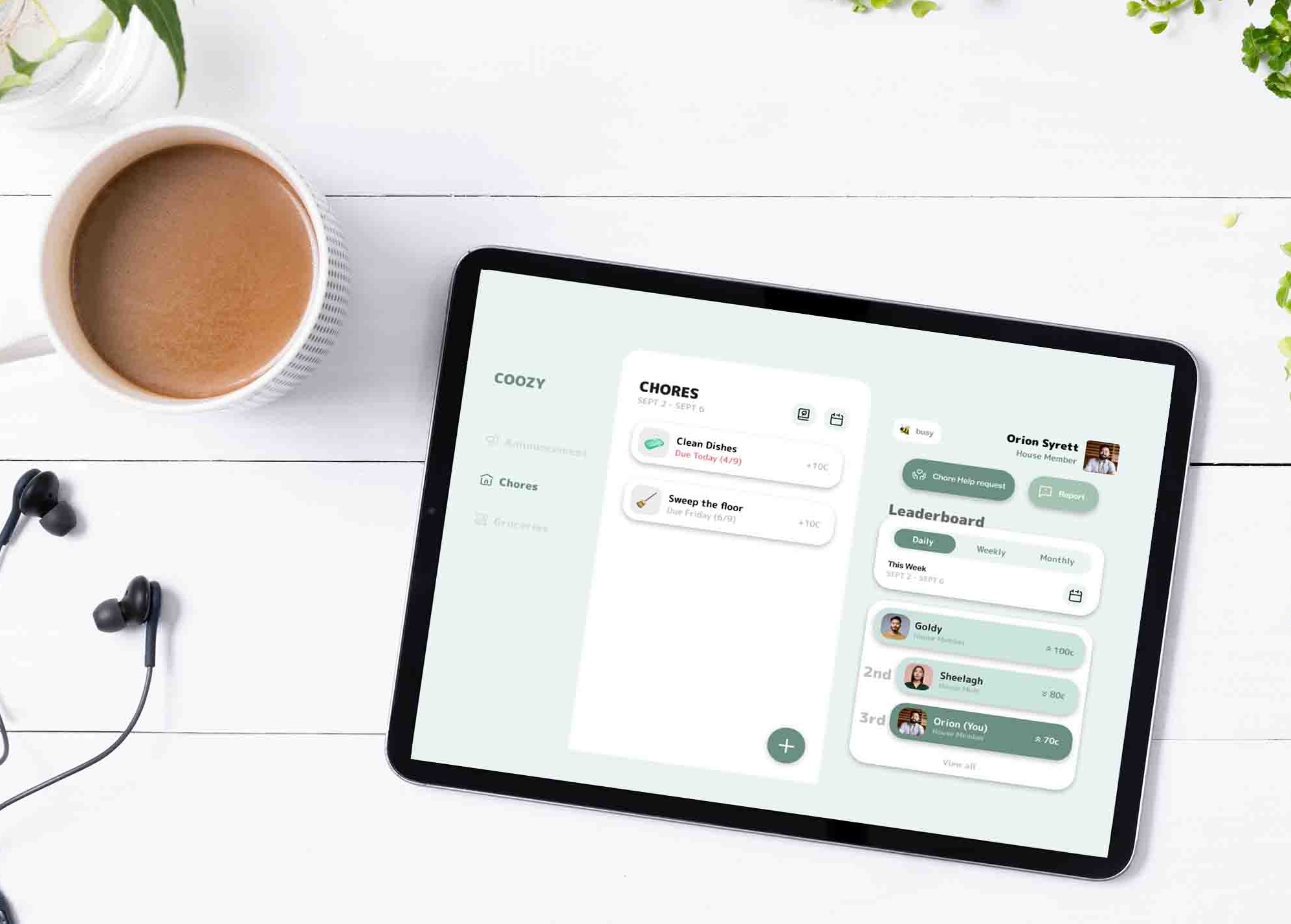 A tablet displays a well-crafted chore app with a task list and leaderboard. Showcasing thoughtful app design, it rests on a white table beside a cup of coffee, earphones, a notepad, and green plant leaves.