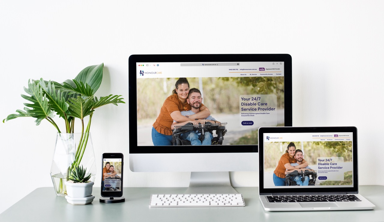 A desktop, laptop and phone on a desk display the same disability care service website (HonourCare) in 3 size formats. A potted plant and notebook sit nearby. The website features a smiling woman hugging a man in a wheelchair.